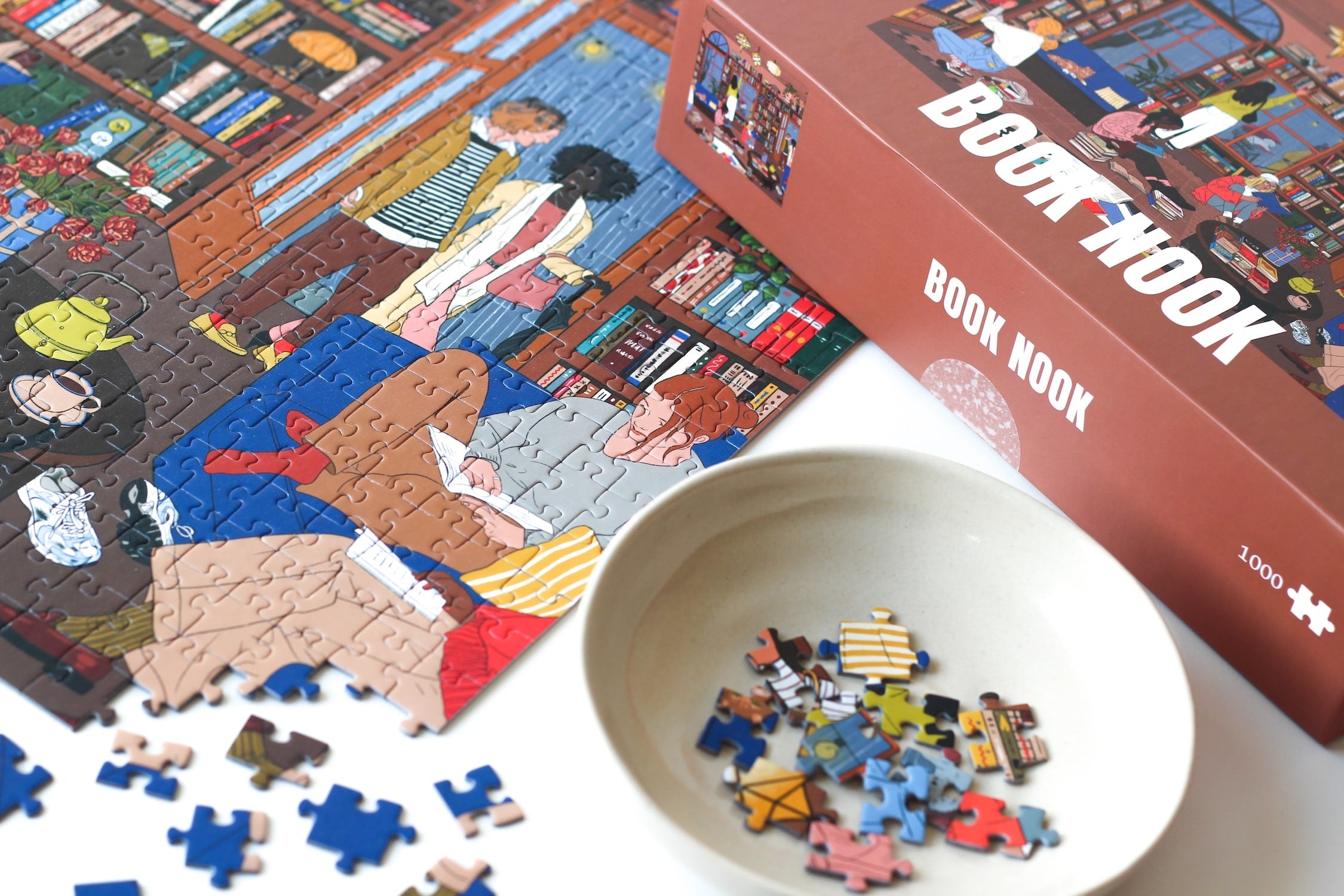 1000-Teile-Puzzle „Book Nook“ von Piecely – Illustration einer gemütlichen Bibliothek mit Bücherregalen, Lesenden und warmem Licht, auf brauner Verpackung.