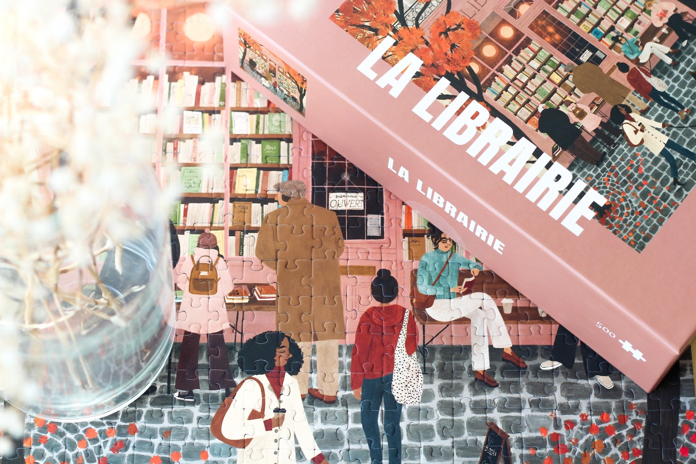 500-Teile-Puzzle „La Librairie“ von Piecely – Illustration einer herbstlichen Pariser Straßenszene vor einer kleinen Buchhandlung, auf rosa-brauner Verpackung.