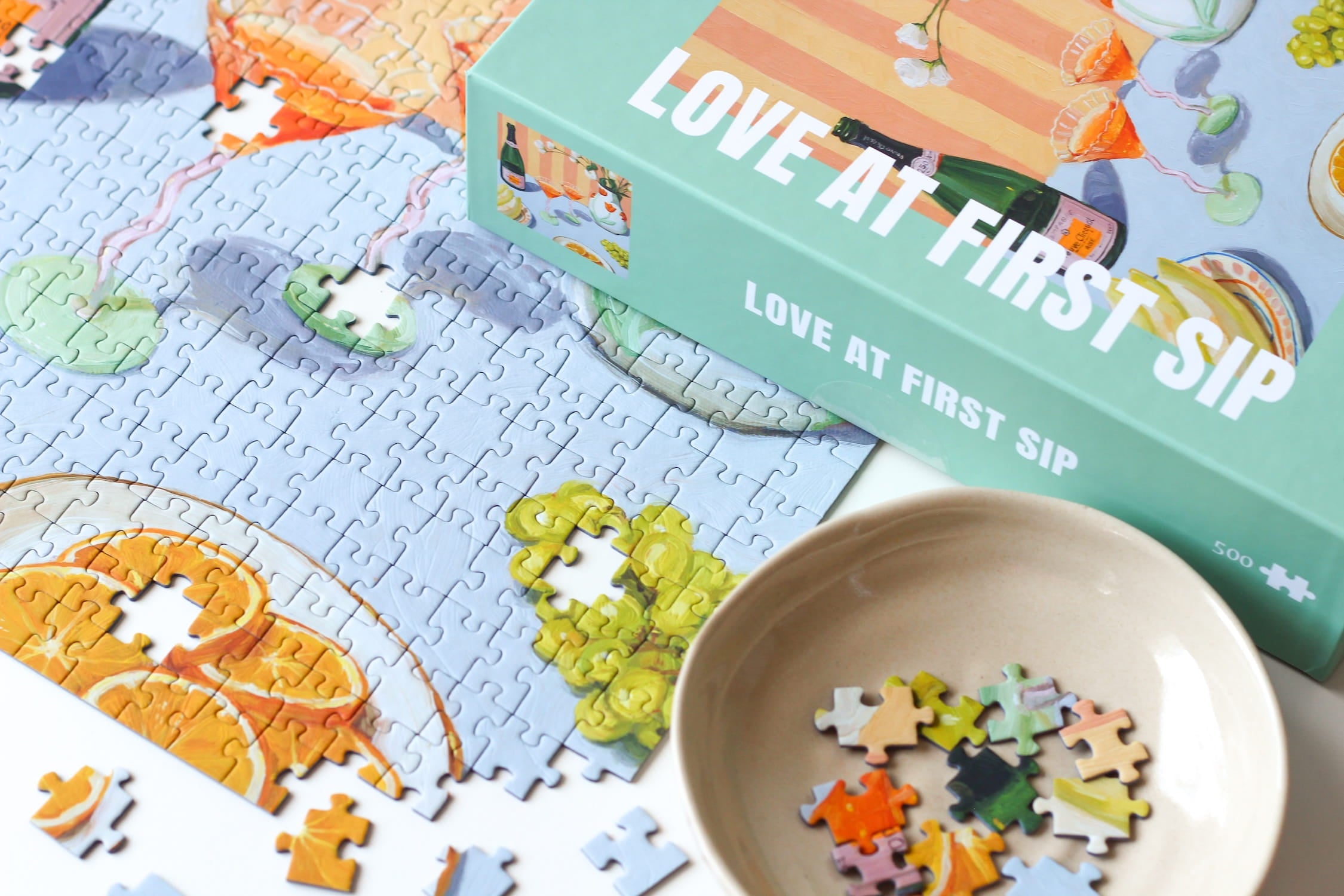 Illustration eines gedeckten Tisches mit Getränken, Orangen, Trauben und Blumen in Vase, als 500-Teile Puzzle ‚Love At First Sip‘ von Piecely