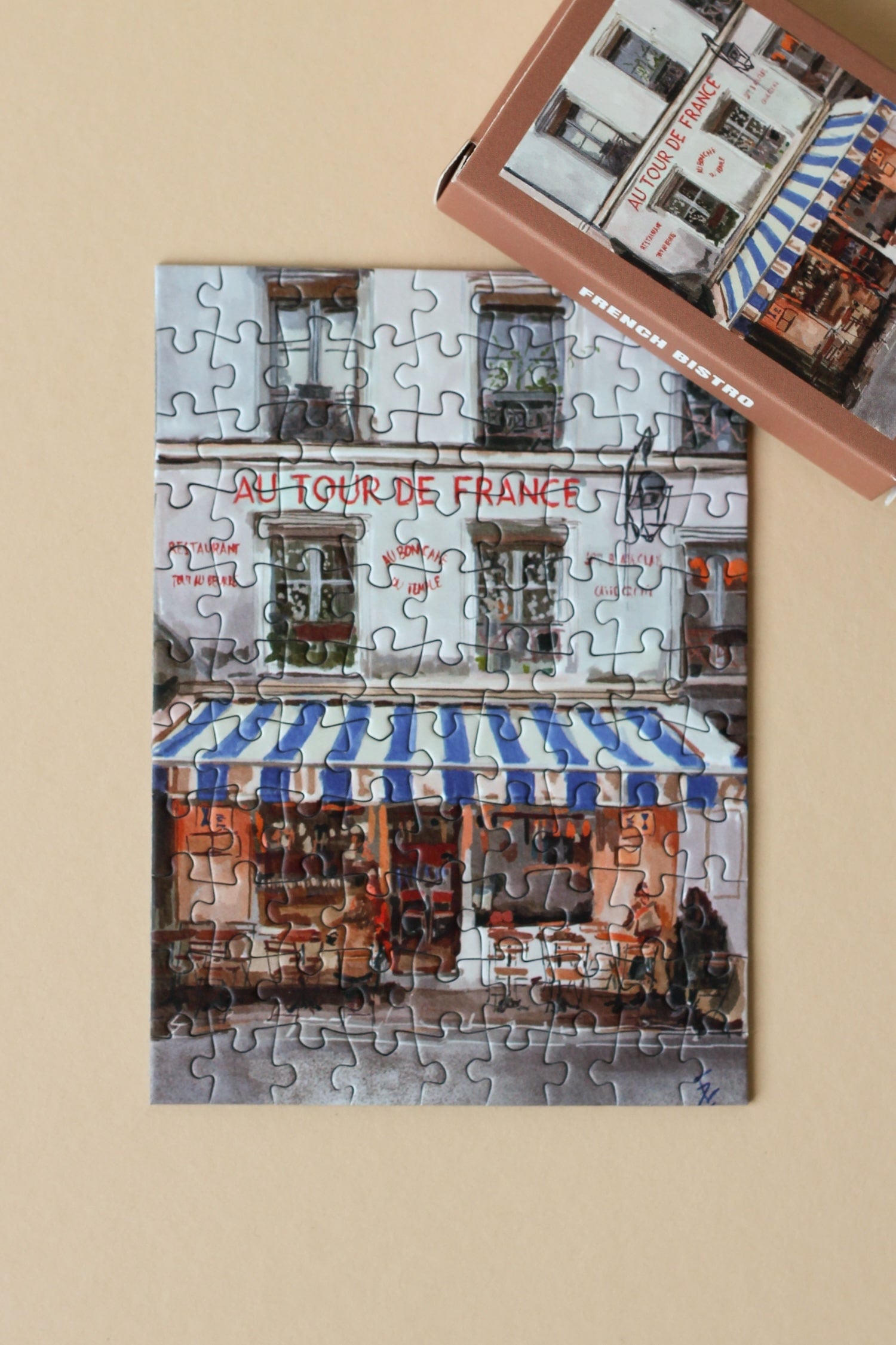 Plastikfreies Minipuzzle für Erwachsene, das ein französisches Bistro mit einer blau-weiß-gestreiften Markise zeigt.