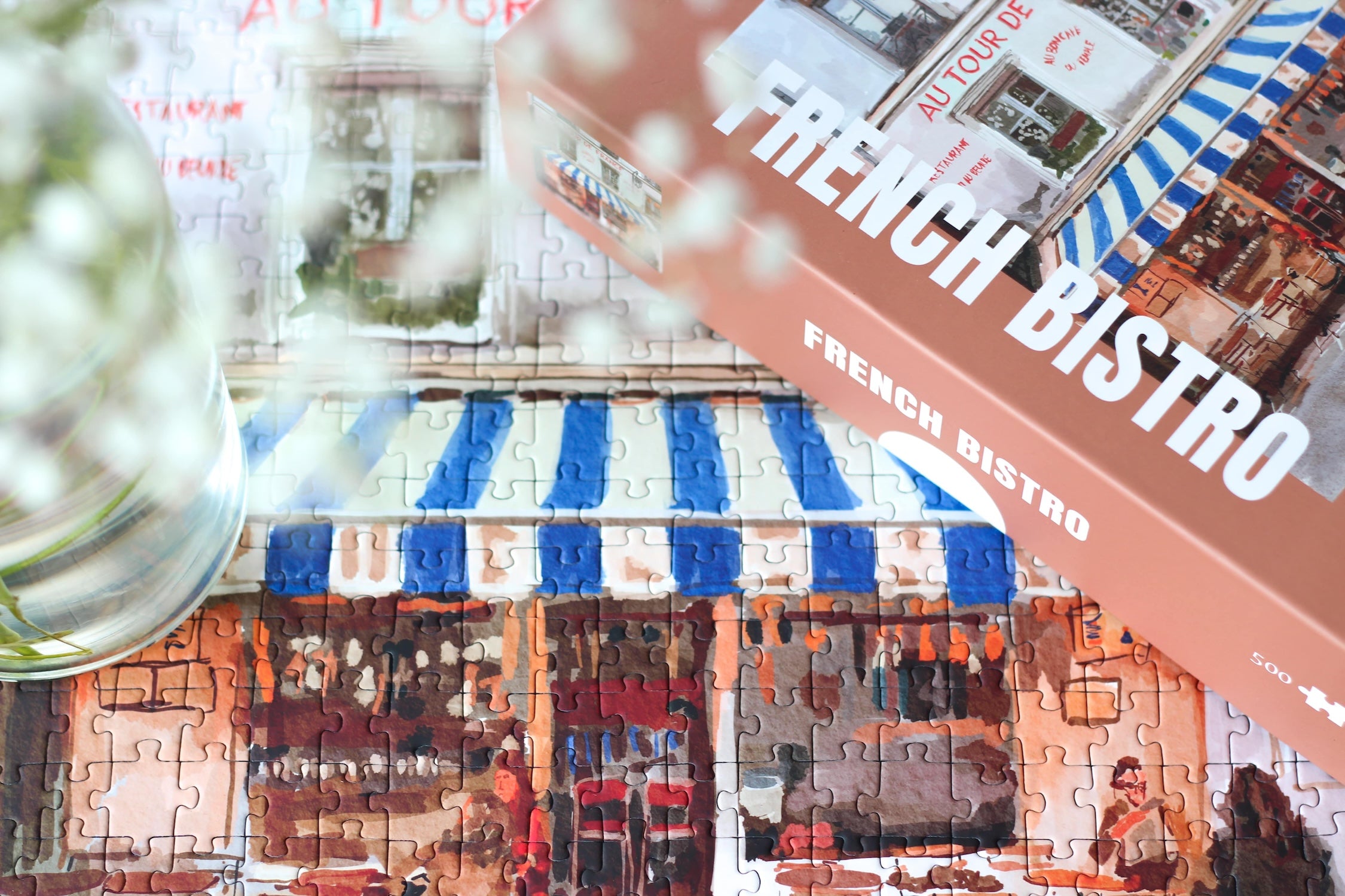 Plastikfreies Puzzle für Erwachsene, das ein französisches Bistro in Paris mit einer blau-weiß-gestreiften Markise zeigt.
