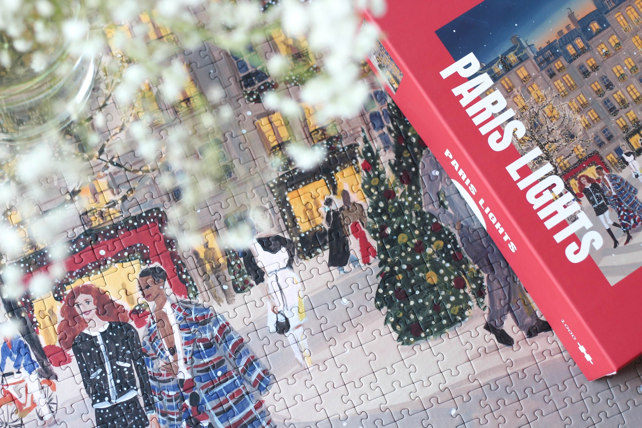 Plastikfreies Puzzle für Erwachsene, das Paris zur Weihnachtszeit zeigt. Stylisch gekleidete Menschen schlendern durch die Straßen und Bäume und Häuser sind mit leuchtenden Lichterketten geschmückt.