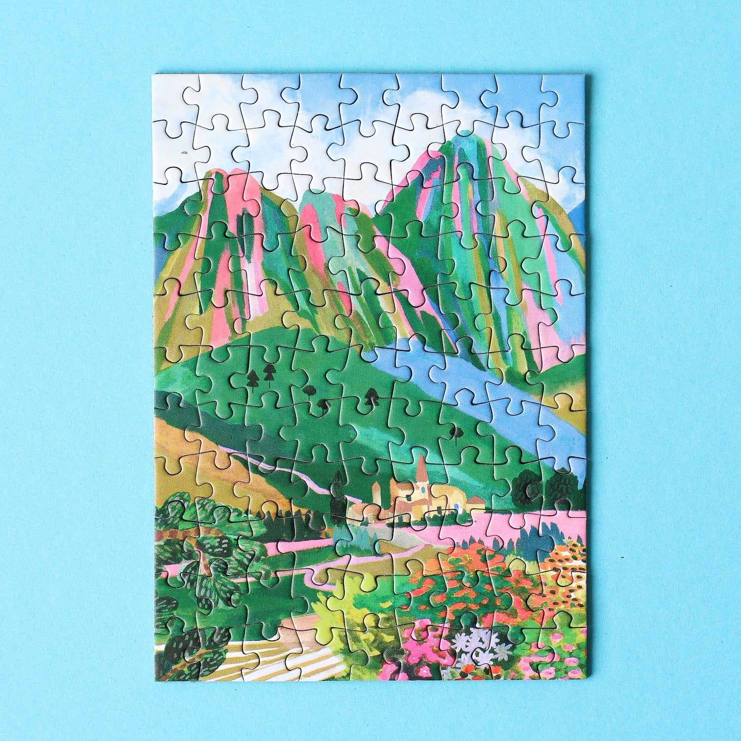 Plastikfreies Minipuzzle für Erwachsene, das die Dolomiten in Italien zeigt. Zu sehen sind ein kleines Dorf, die Berge und ein buntes Blumenmeer