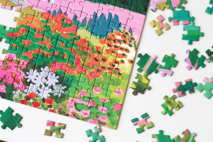 1000-Teile-Puzzle „The Dolomites“ von Piecely – Illustration einer farbenfrohen Berglandschaft in den Dolomiten mit Dorf und Blumen, auf türkisfarbener Verpackung.
