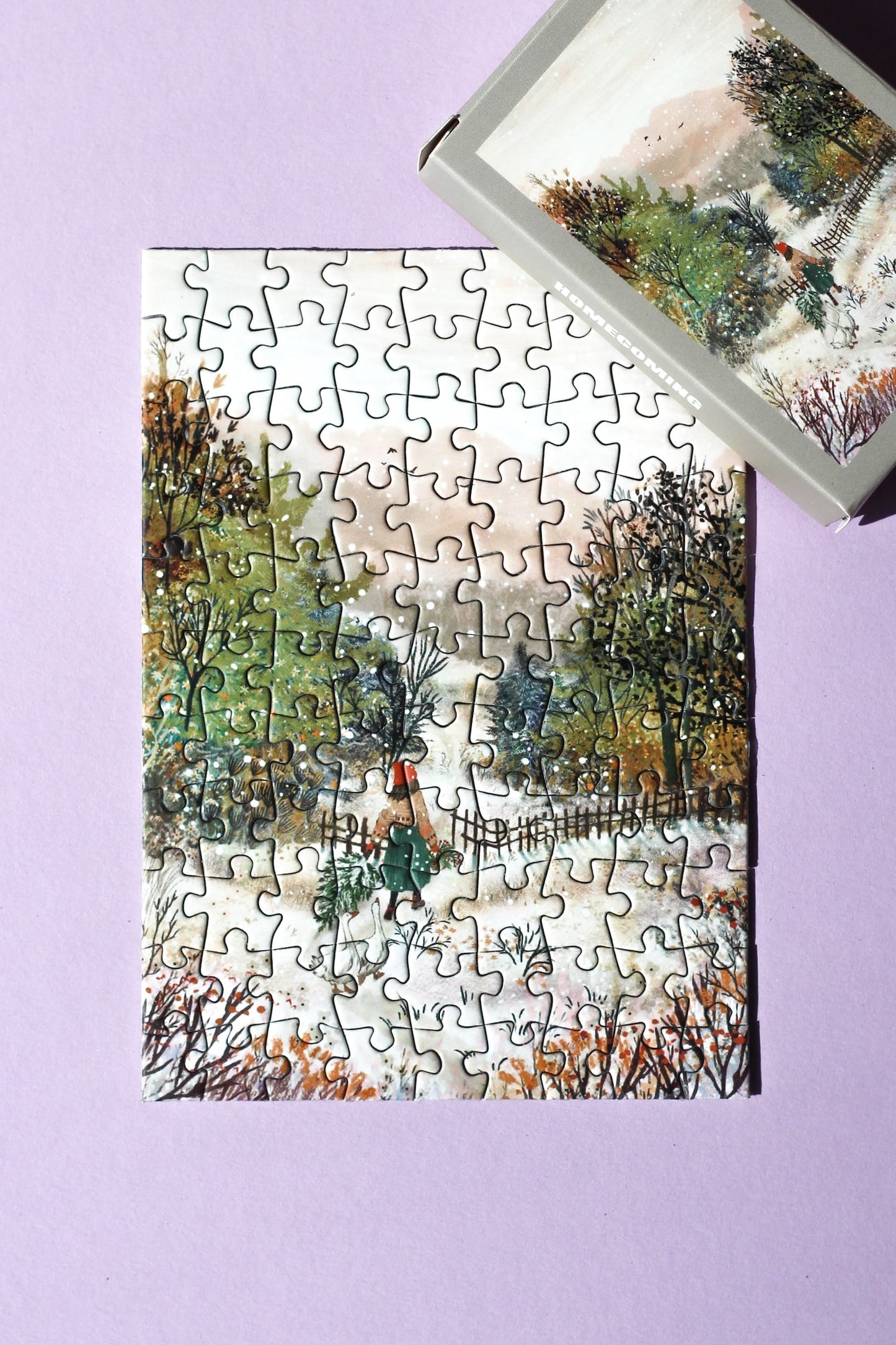 99-Teile-Mini-Puzzle „Homecoming“ von Piecely – Illustration einer winterlichen Landschaft mit Frau, zwei Gänsen und Weihnachtsbaum, auf grauer Verpackung