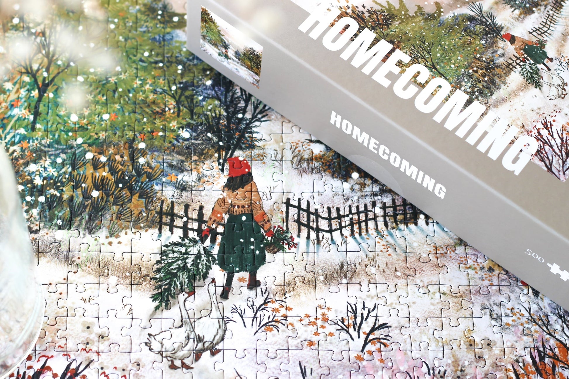 500-Teile-Puzzle „Homecoming“ von Piecely – Illustration einer winterlichen Landschaft mit Frau und zwei Gänsen auf einem verschneiten Weg, auf grauer Verpackung.