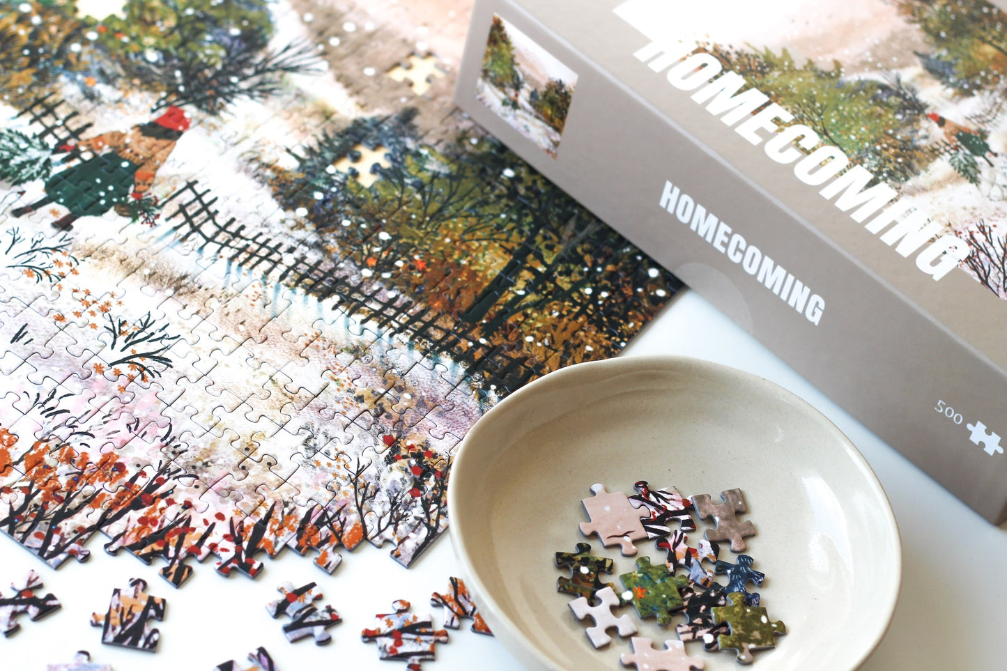 500-Teile-Puzzle „Homecoming“ von Piecely – Illustration einer winterlichen Landschaft mit Frau und zwei Gänsen auf einem verschneiten Weg, auf grauer Verpackung.