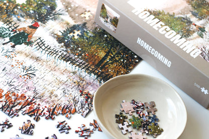 500-Teile-Puzzle „Homecoming“ von Piecely – Illustration einer winterlichen Landschaft mit Frau und zwei Gänsen auf einem verschneiten Weg, auf grauer Verpackung.