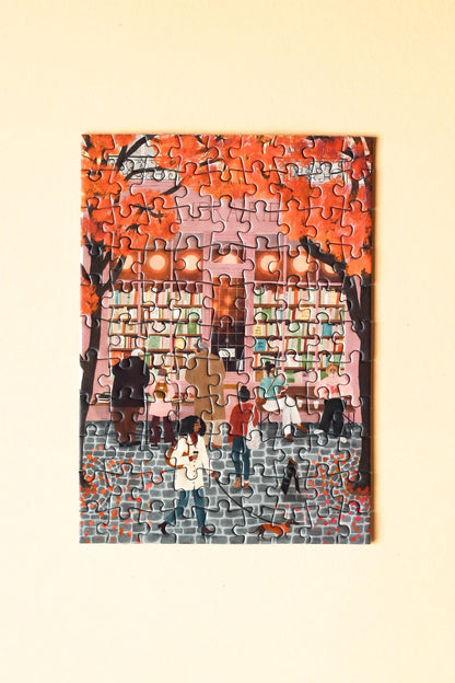 99-Teile-Mini-Puzzle „La Librairie“ von Piecely – Illustration einer herbstlichen Straßenszene vor einer Pariser Buchhandlung, auf rosafarbener Verpackung.