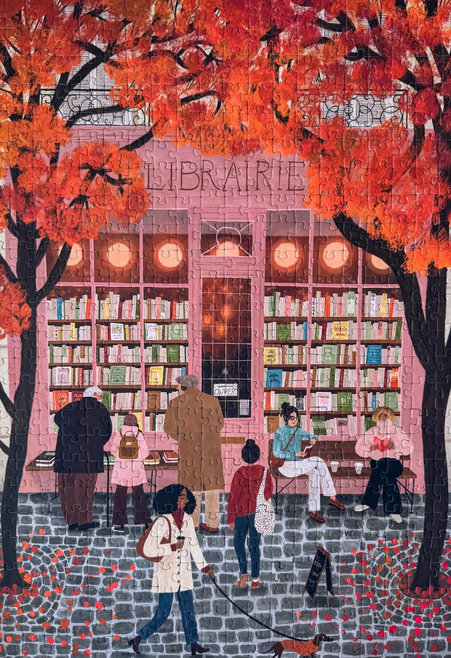500-Teile-Puzzle „La Librairie“ von Piecely – Illustration einer herbstlichen Pariser Straßenszene vor einer kleinen Buchhandlung, auf rosa-brauner Verpackung.