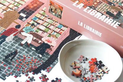 500-Teile-Puzzle „La Librairie“ von Piecely – Illustration einer herbstlichen Pariser Straßenszene vor einer kleinen Buchhandlung, auf rosa-brauner Verpackung.