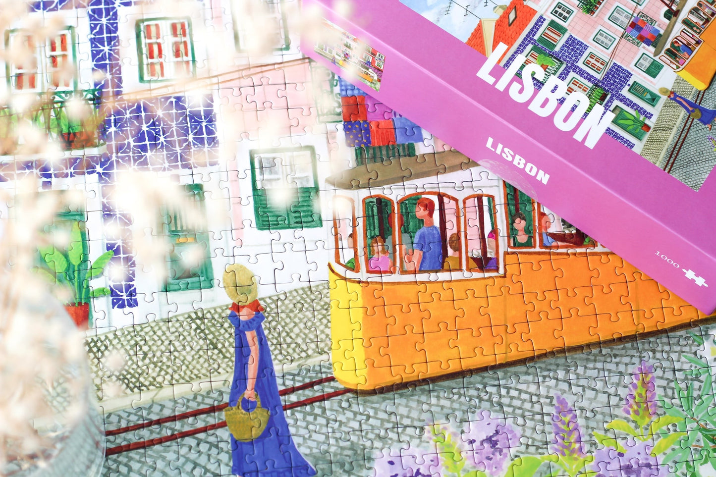 1000-Teile-Puzzle „Lisbon“ von Piecely – Illustration einer farbenfrohen Straßenszene in Lissabon mit gelber Straßenbahn und bunten Häusern, auf pinker Verpackung.