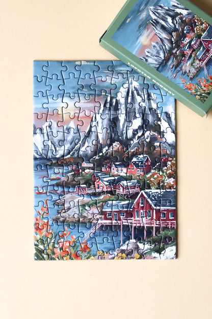 99-Teile-Mini-Puzzle „Lofoten Norway“ von Piecely – Illustration einer norwegischen Küstenlandschaft mit Bergen, roten Häusern und Meer, auf olivgrüner Verpackung.