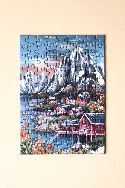 99-Teile-Mini-Puzzle „Lofoten Norway“ von Piecely – Illustration einer norwegischen Küstenlandschaft mit Bergen, roten Häusern und Meer, auf olivgrüner Verpackung.