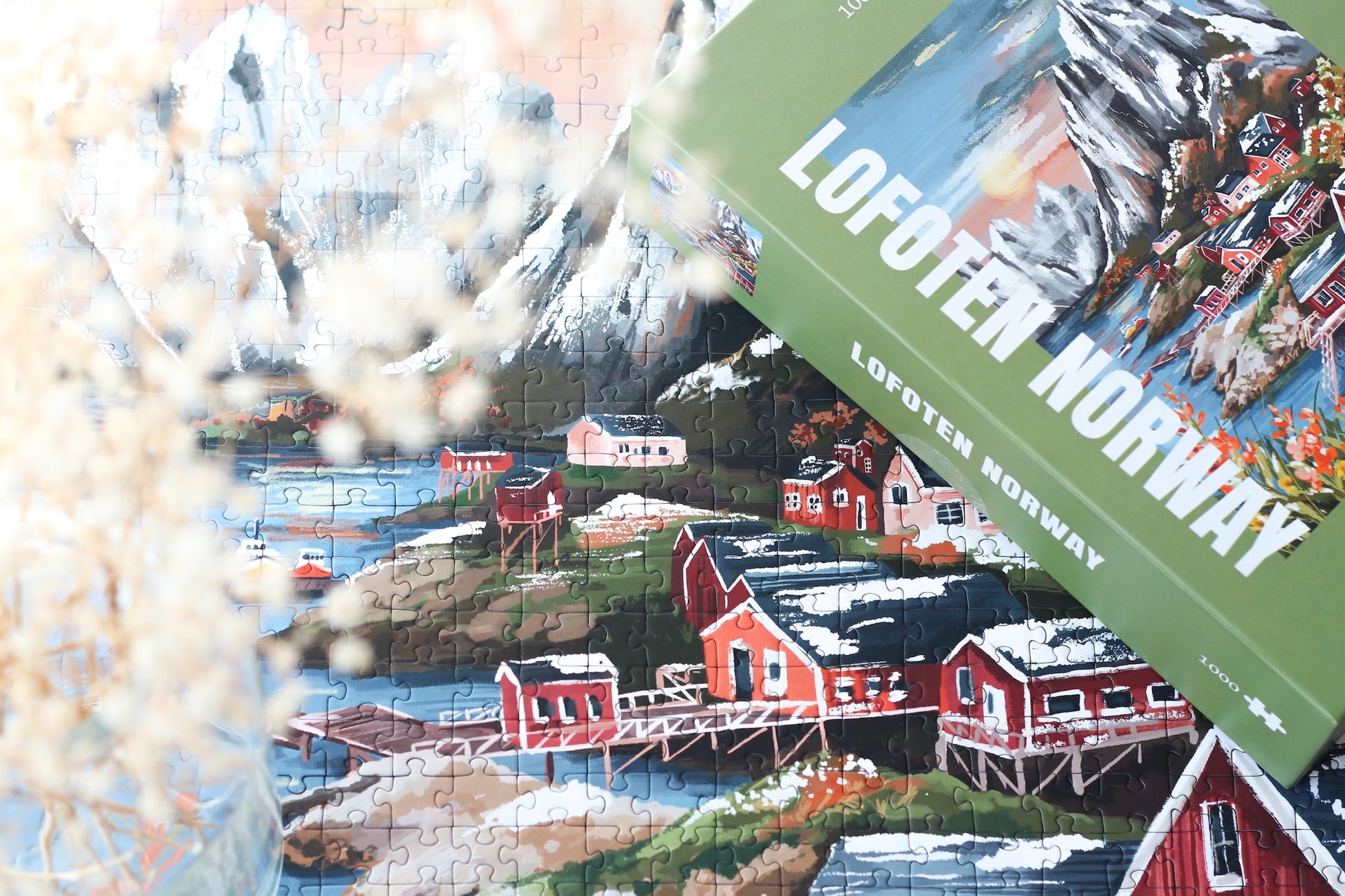 1000-Teile-Puzzle „Lofoten Norway“ von Piecely – Illustration einer norwegischen Küstenlandschaft mit schneebedeckten Bergen, roten Holzhäusern und Meer, auf olivgrüner Verpackung.