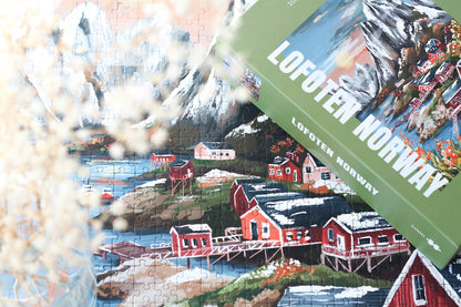 1000-Teile-Puzzle „Lofoten Norway“ von Piecely – Illustration einer norwegischen Küstenlandschaft mit schneebedeckten Bergen, roten Holzhäusern und Meer, auf olivgrüner Verpackung.