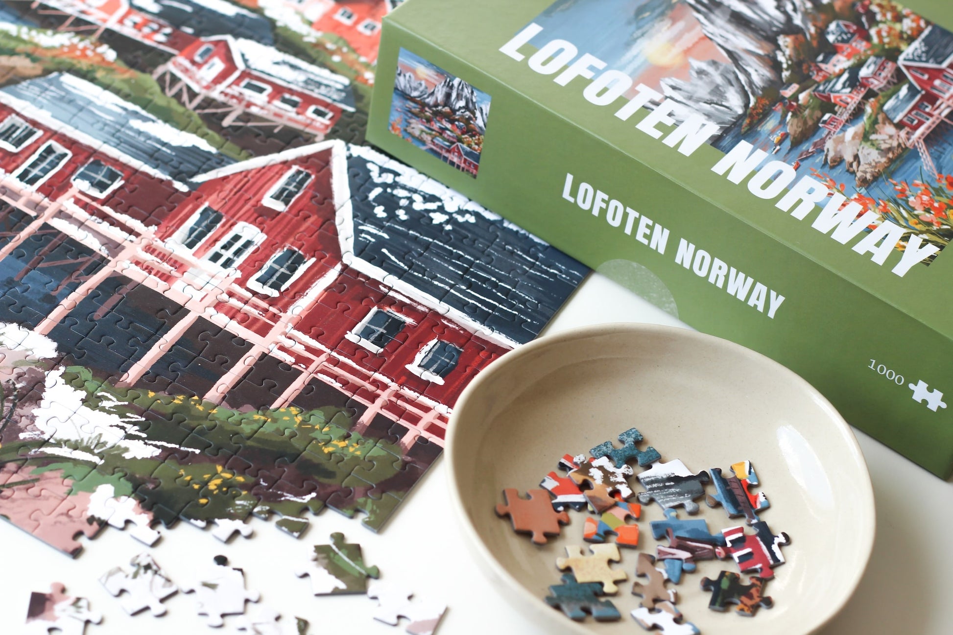 1000-Teile-Puzzle „Lofoten Norway“ von Piecely – Illustration einer norwegischen Küstenlandschaft mit schneebedeckten Bergen, roten Holzhäusern und Meer, auf olivgrüner Verpackung.