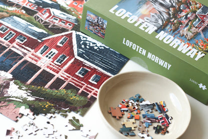 1000-Teile-Puzzle „Lofoten Norway“ von Piecely – Illustration einer norwegischen Küstenlandschaft mit schneebedeckten Bergen, roten Holzhäusern und Meer, auf olivgrüner Verpackung.