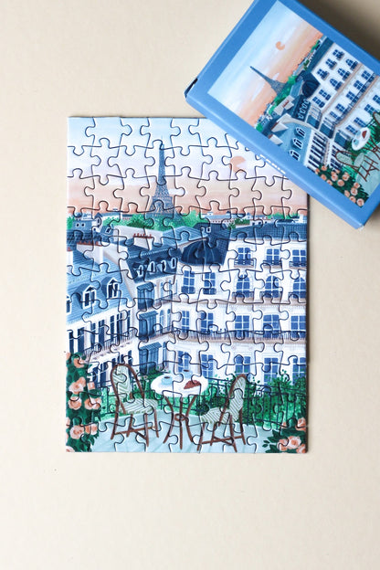 99-Teile-Mini-Puzzle „Paris“ von Piecely – Illustration eines Pariser Balkons mit Frühstückstisch, Rosen und Blick auf den Eiffelturm, auf hellblauer Verpackung.