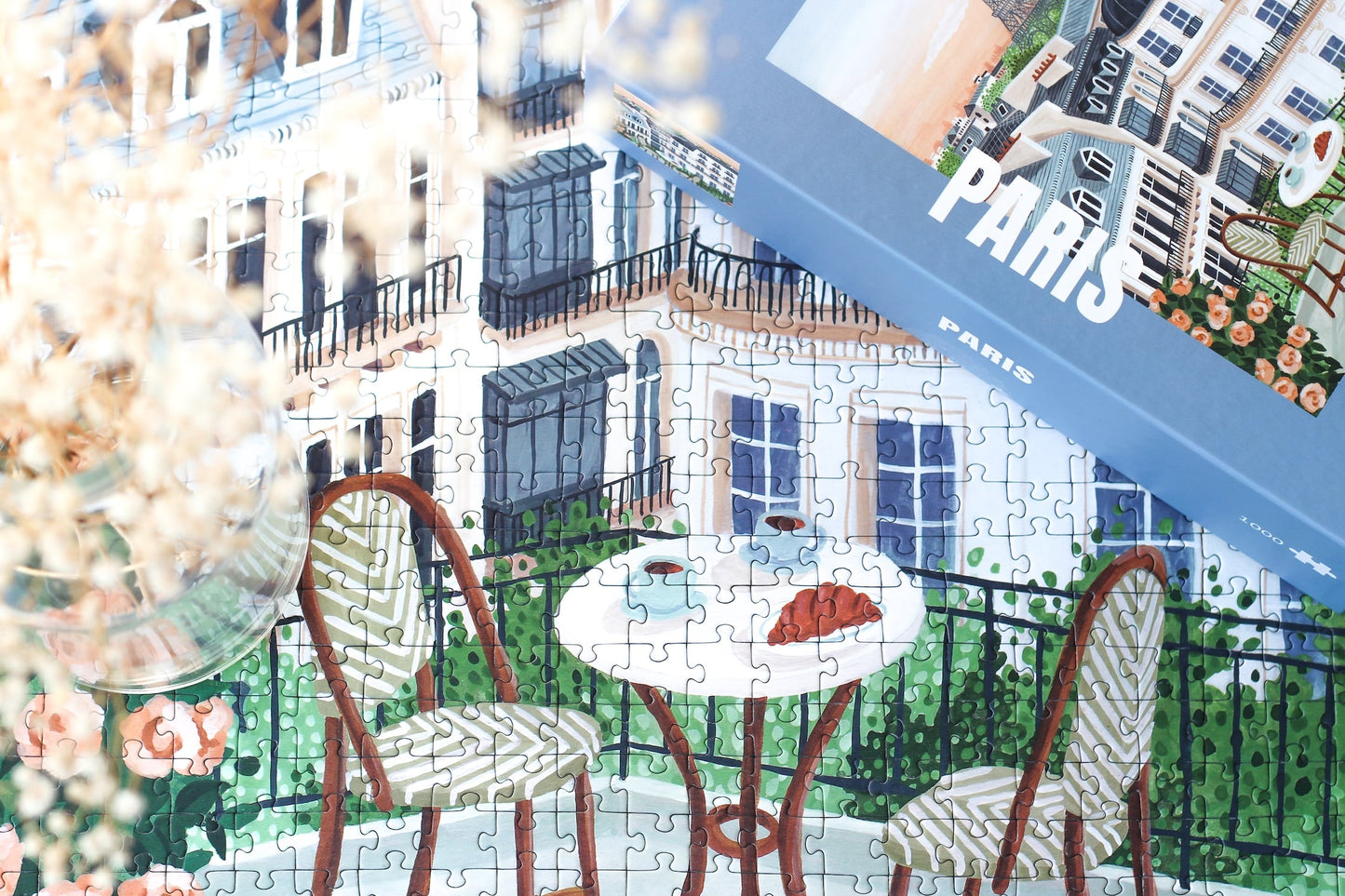 1000-Teile-Puzzle „Paris“ von Piecely – Illustration eines Pariser Balkons mit Frühstückstisch und Blick auf den Eiffelturm, auf hellblauer Verpackung.