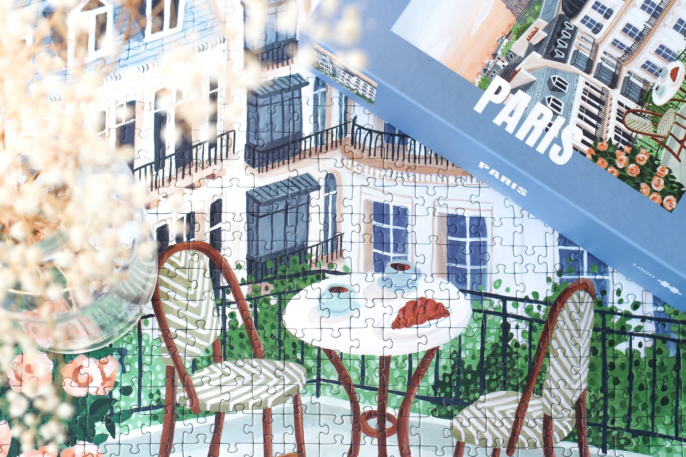 1000-Teile-Puzzle „Paris“ von Piecely – Illustration eines Pariser Balkons mit Frühstückstisch und Blick auf den Eiffelturm, auf hellblauer Verpackung.