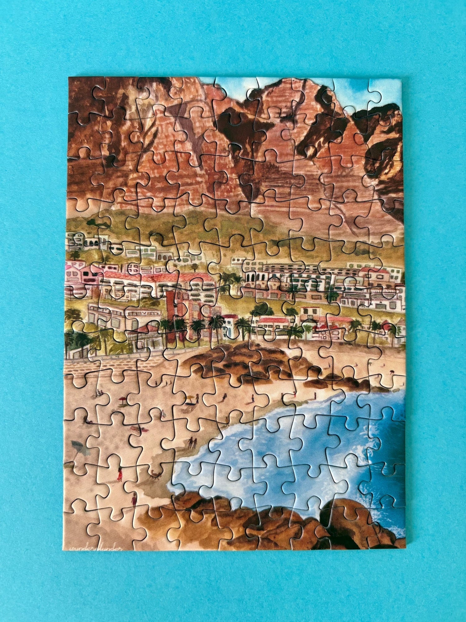 99-teiliges Minipuzzle für Erwachsene, das die Strandpromenade des Kapstädter Viertels Camps Bay zeigt. Im Vordergrund sind man Sandstrand und blaues Meer und im Hintergrund Häuserketten und die Berge.