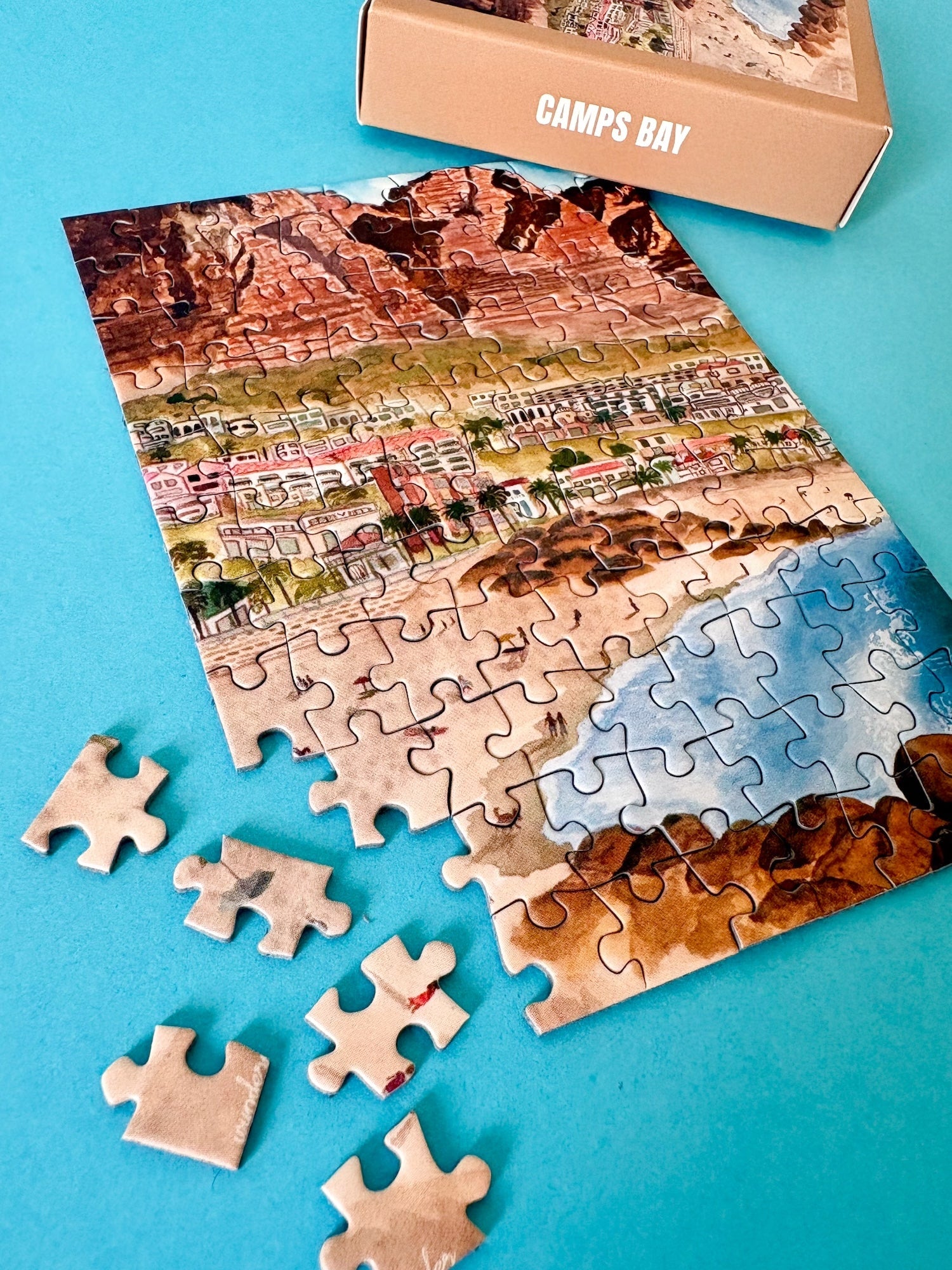 99-teiliges Minipuzzle für Erwachsene, das die Strandpromenade des Kapstädter Viertels Camps Bay zeigt. Im Vordergrund sind man Sandstrand und blaues Meer und im Hintergrund Häuserketten und die Berge.