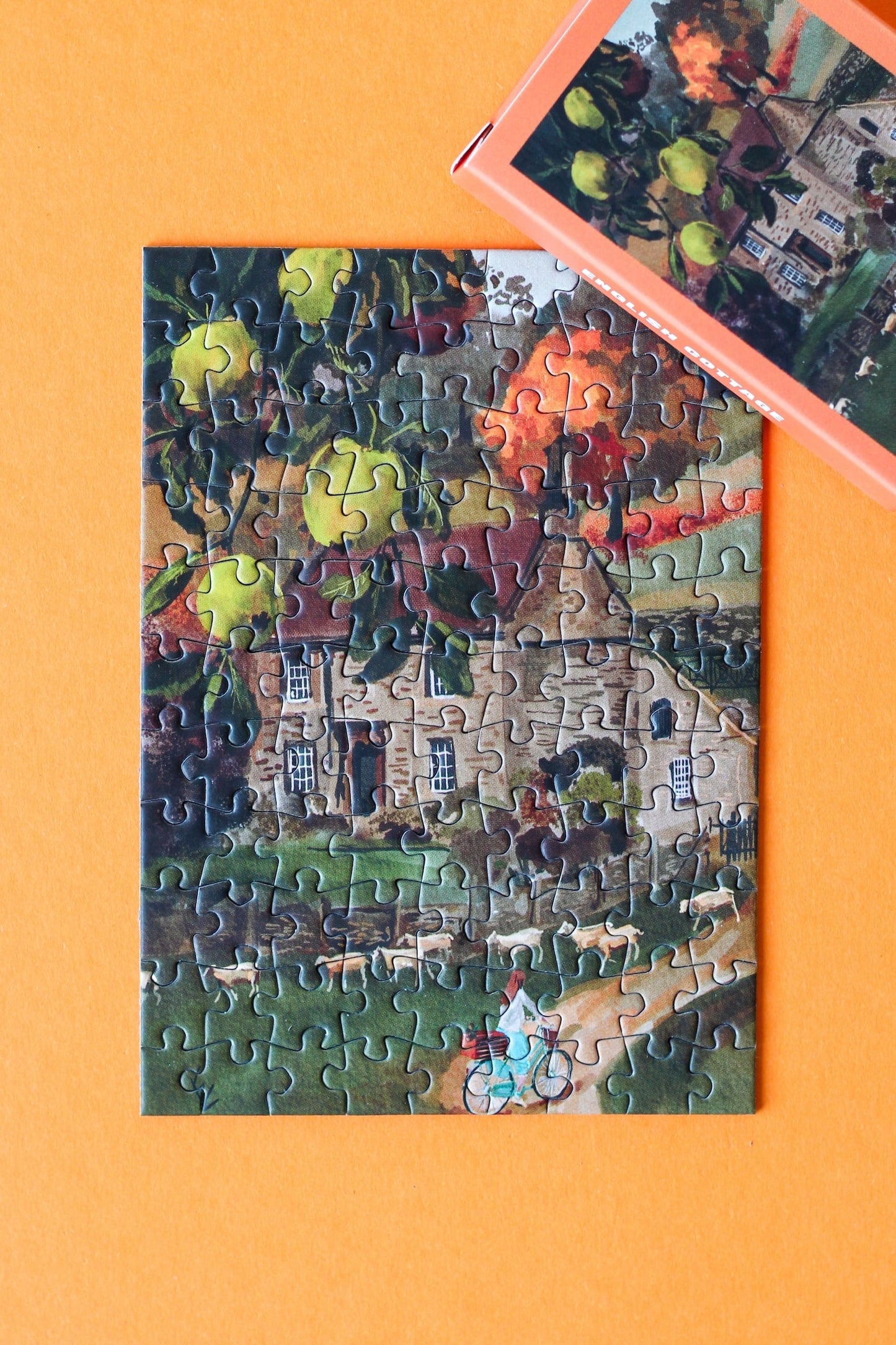 Plastikfreies Minipuzzle für Erwachsene, das ein Cottage auf dem englischen Land im Herbst zeigt. Vor dem Cottage stehen ein Birnbaum und eine Schafherde. Eine Frau fährt auf einem Fahrrad den Weg zum Cottage hoch.