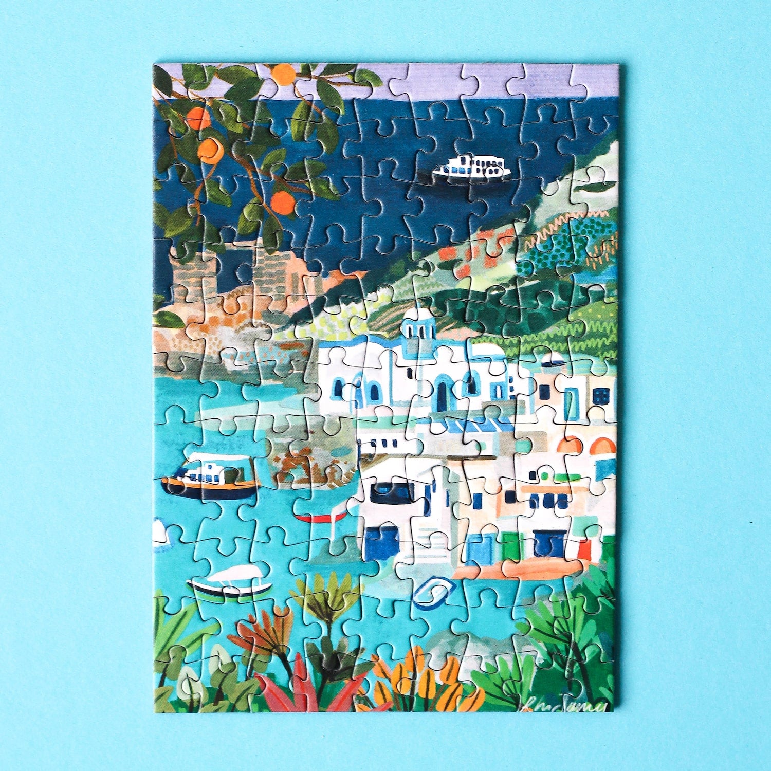 Plastikfreies Minipuzzle, das die Küste der Insel Milos in Griechenland zeigt. Zu sehen sind weiße Häuser, blaues Meer, saftige Orangen und Schiffe
