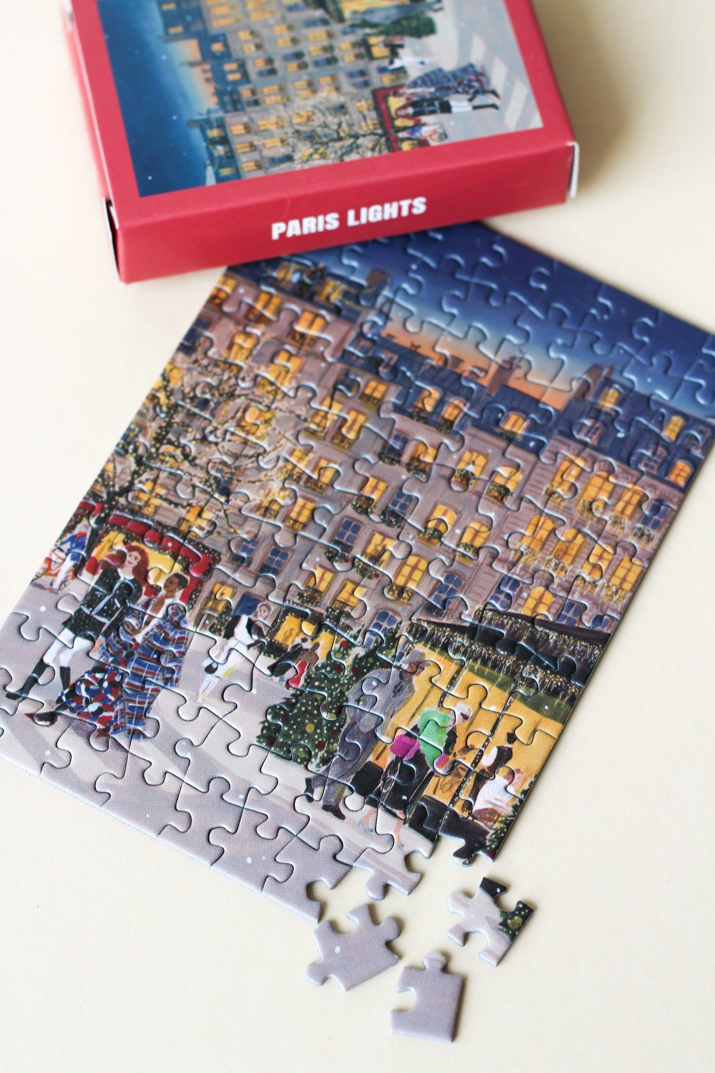 Plastikfreies Minipuzzle für Erwachsene, das Paris zur Weihnachtszeit  zeigt. Stylisch gekleidete Menschen schlendern durch die Straßen und Bäume und Häuser sind mit leuchtenden Lichterketten geschmückt.