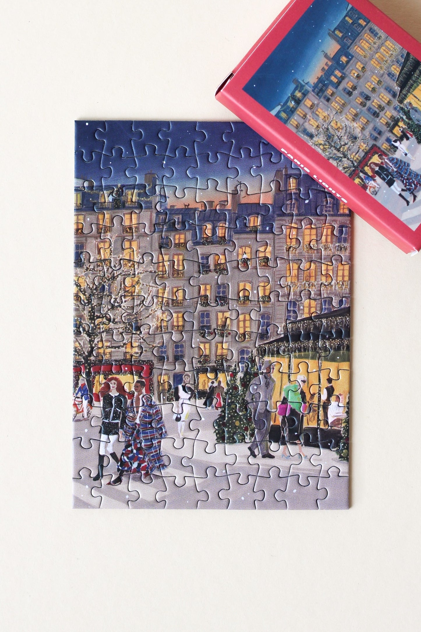 Plastikfreies Minipuzzle für Erwachsene, das Paris zur Weihnachtszeit  zeigt. Stylisch gekleidete Menschen schlendern durch die Straßen und Bäume und Häuser sind mit leuchtenden Lichterketten geschmückt.