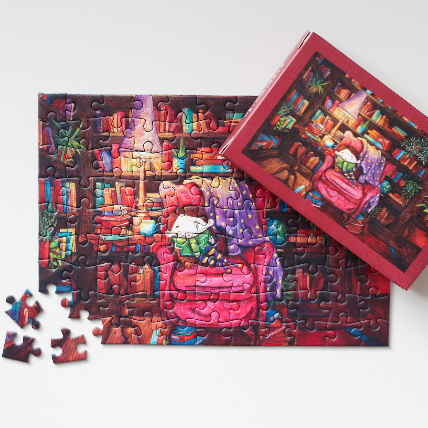 99-teiliges Portable Magic Minipuzzle für Erwachsene, das ein kleines Mädchen zeigt, wie es umgeben von prall gefüllten Bücherregalen auf einem Sessel sitzt und ein Buch liest.