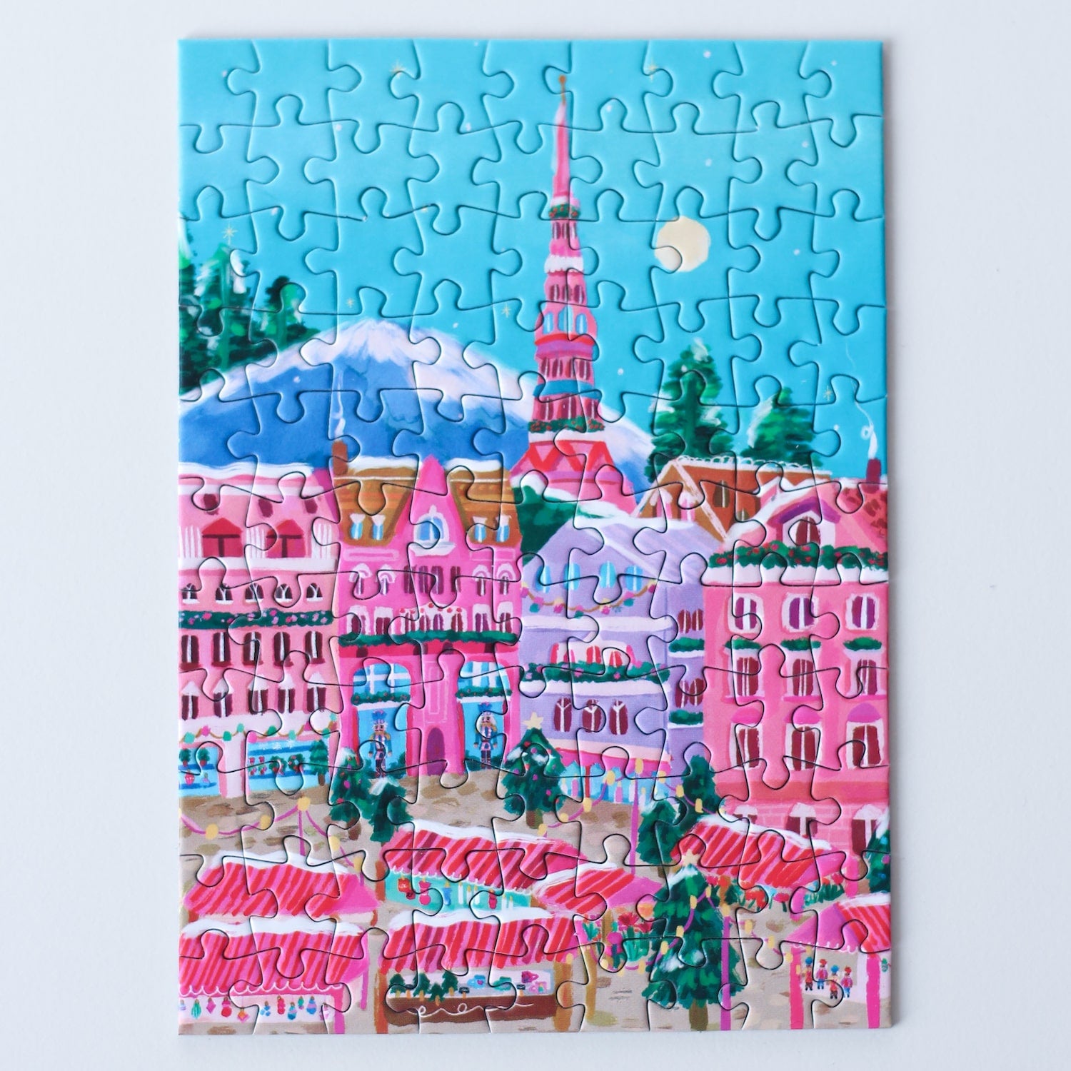 99-teiliges Minipuzzle für Erwachsene, das den Weihnachtsmarkt in Riga zeigt. Auf dem Platz befinden sich pinke Stände und geschmückte Weihnachtsbäume.