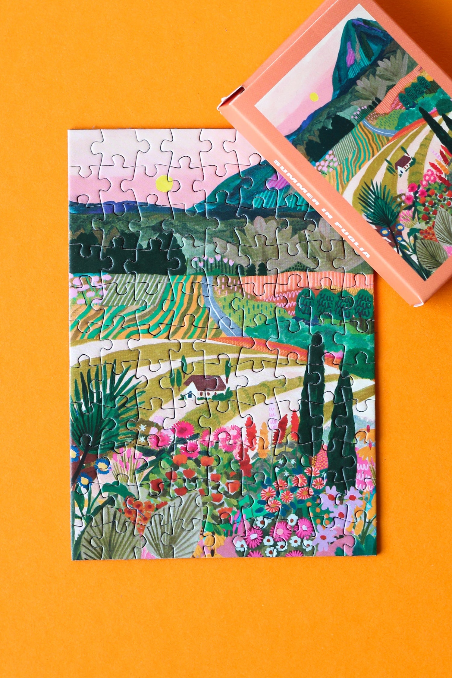 Plastikfreies Minipuzzle für Erwachsene, das die bunte Landschafts Apuliens in Italien zeigt. Zu sehen sind ein Haus, die Berge, bunte Blumen, Felder und die untergehende Sonne