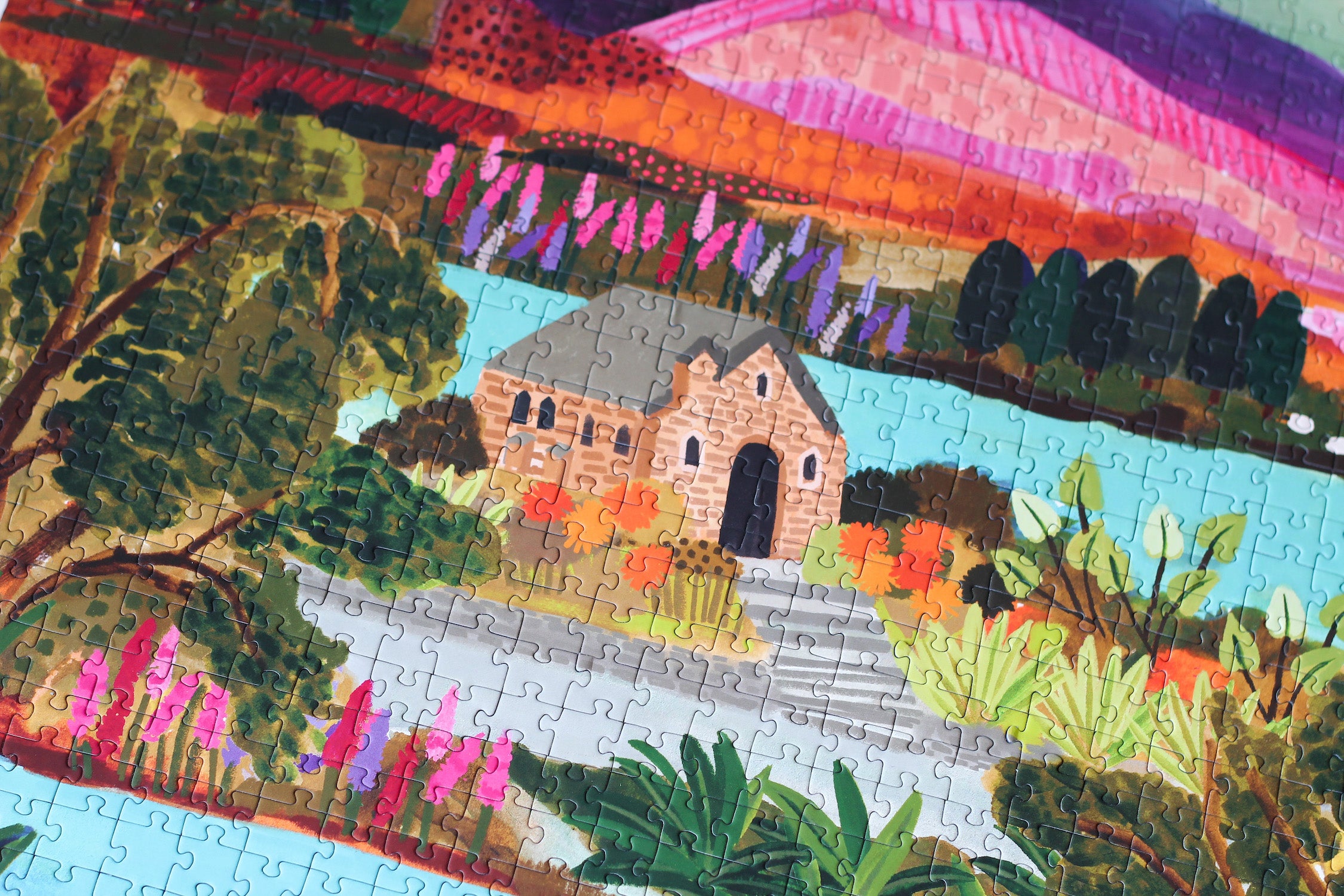 Nachhaltiges Puzzle mit 1000 Teilen, das den Lake Tekapo mit einem Häuschen, den Bergen im Hintergrund und vielen Pflanzen und Blumen zeigt.