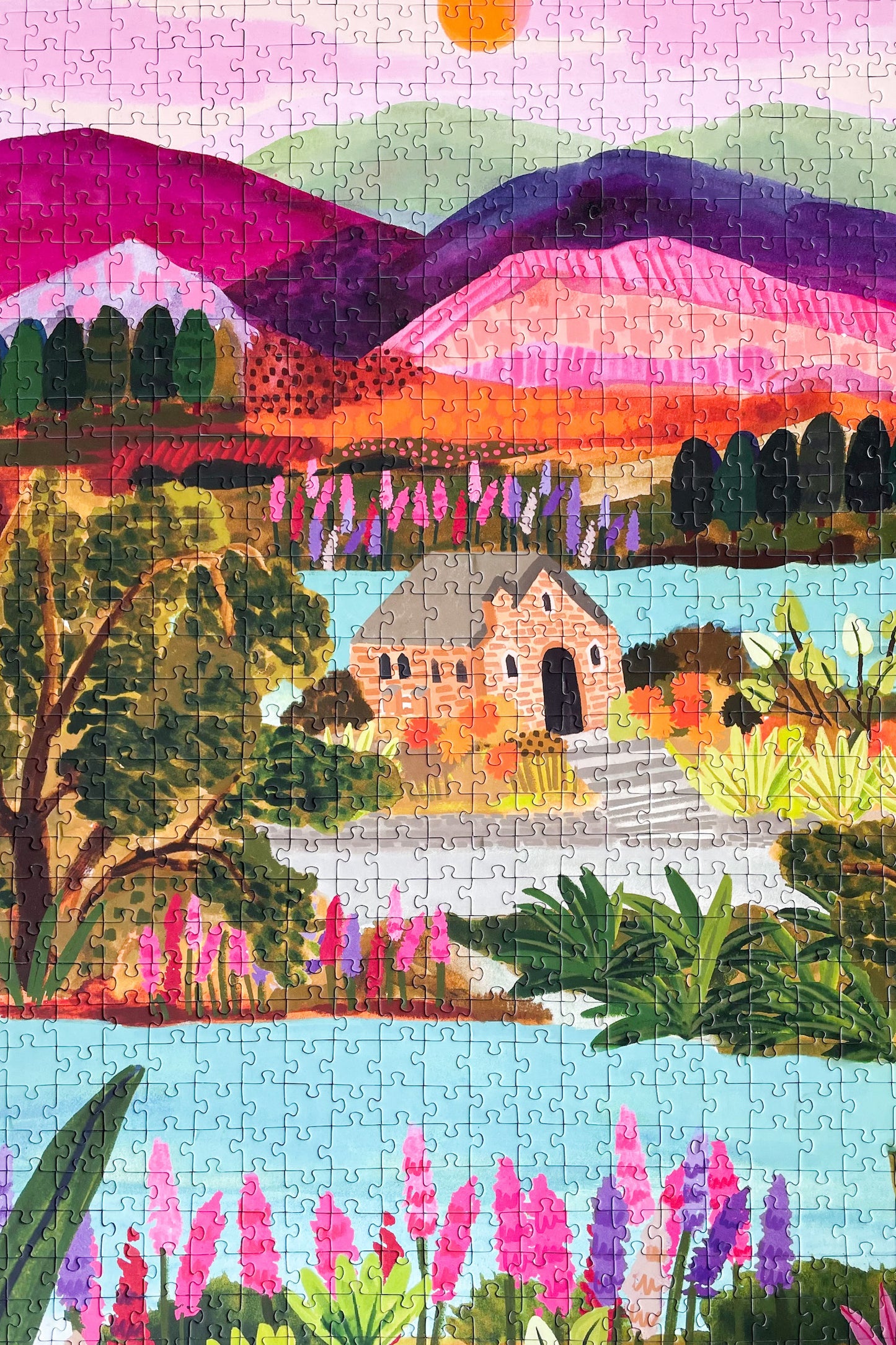 Nachhaltiges Puzzle mit 1000 Teilen, das den Lake Tekapo mit einem Häuschen, den Bergen im Hintergrund und vielen Pflanzen und Blumen zeigt.